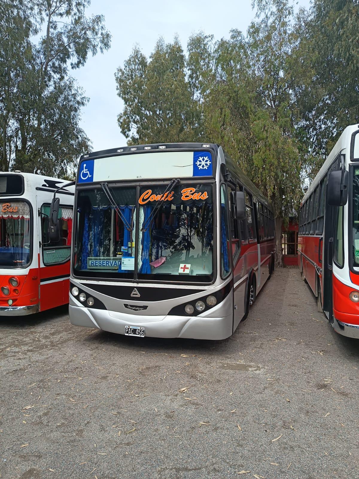 Frente de un micro gris de Conti Bus con el nombre de la empresa en el parabrisas, estacionado entre árboles junto a otros micros